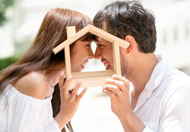 Casal sorridente olhando um para o outro através de uma estrutura de madeira em forma de casa, simbolizando planos de habitação.